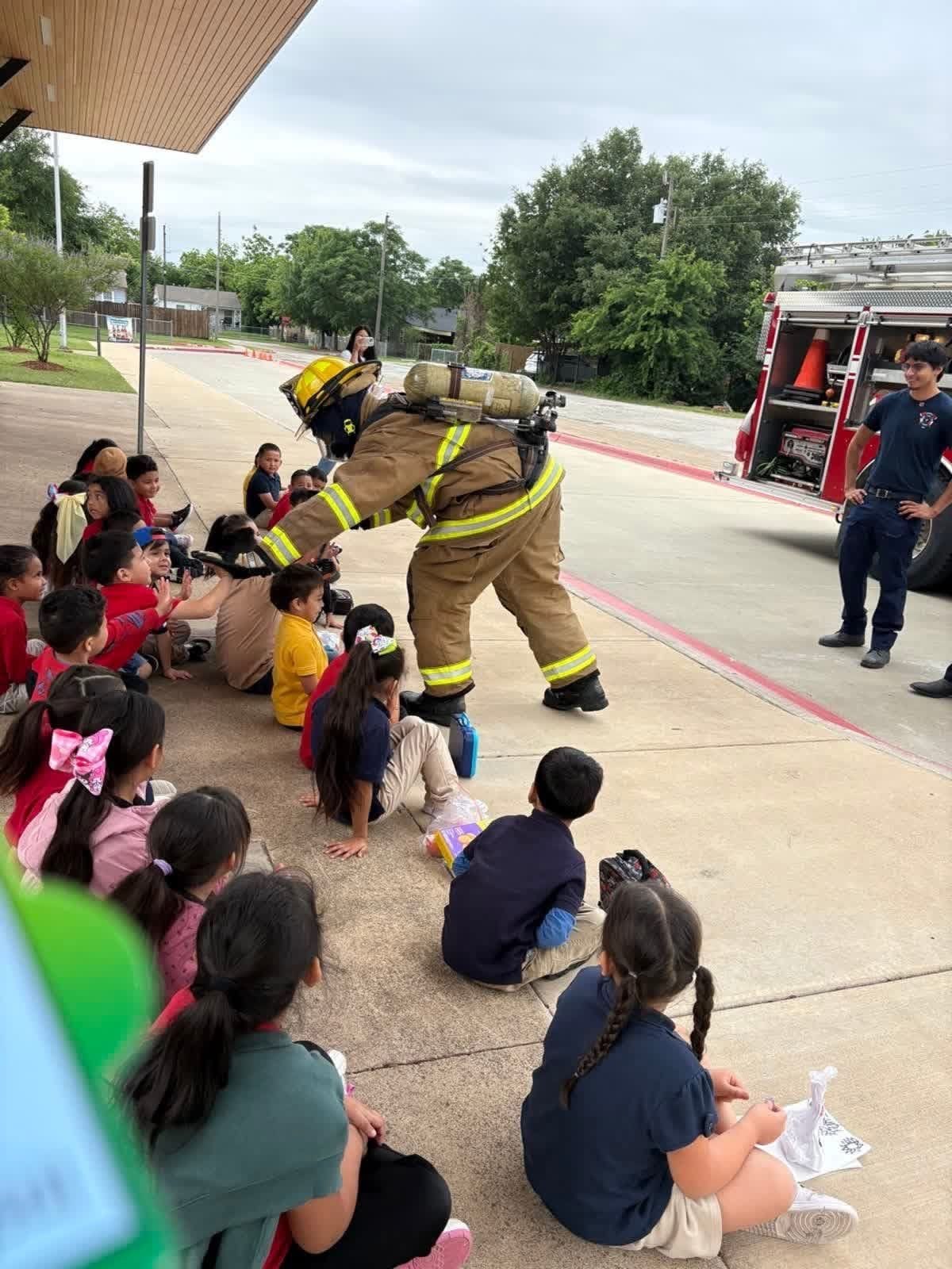 A firefighter in full gear and air pack shows a group of seated schoolchildren what a firefighter looks like on the job